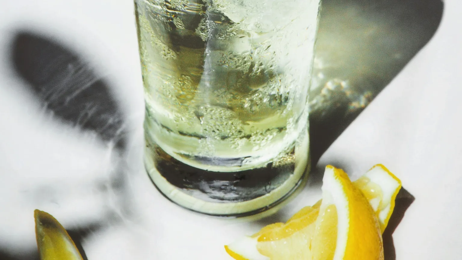 A tall glass of chilled sparkling drink over ice with a lemon wedge in bright sunlight