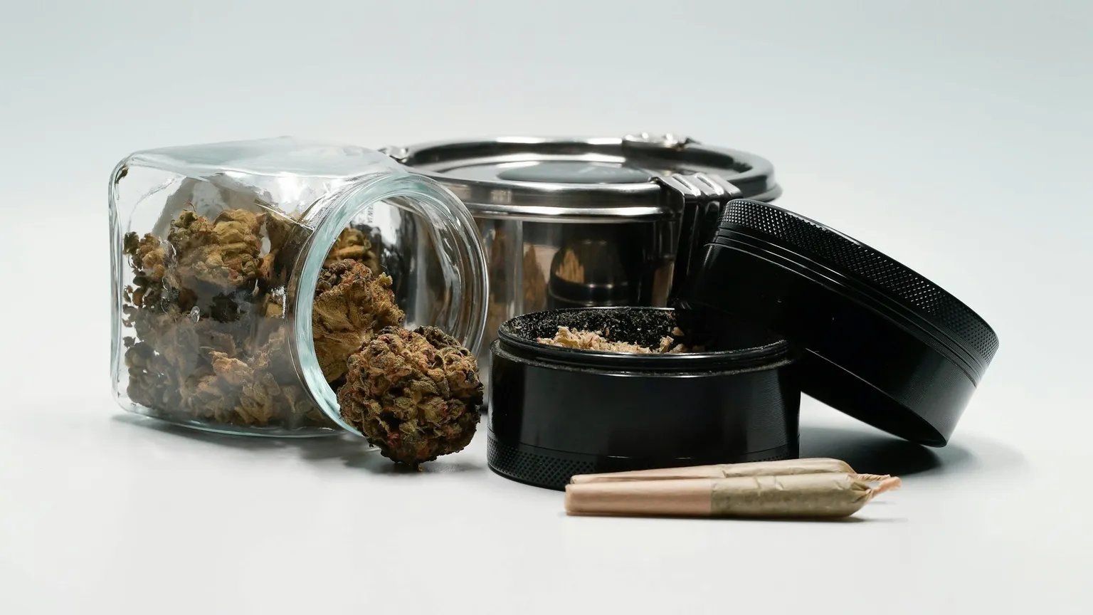 A glass jar of cannabis flower beside a grinder and two pre-rolls on a plain background