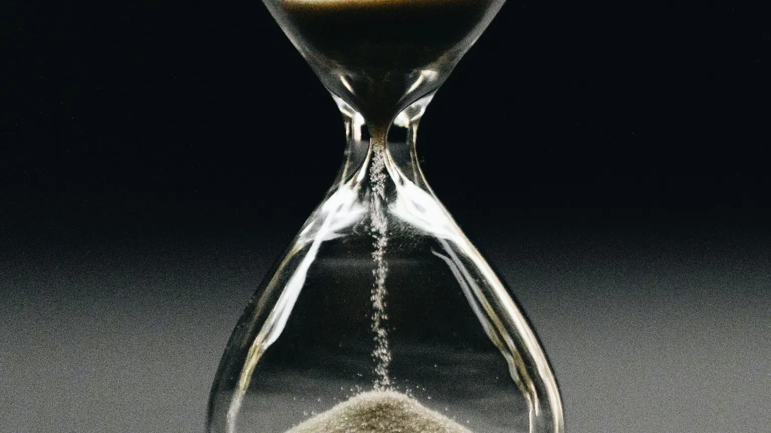 Sand falling through the narrow neck of a glass hourglass against a dark background