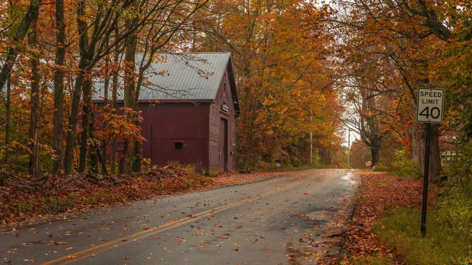 A quiet New England country road past a red barn under autumn foliage on a rainy day