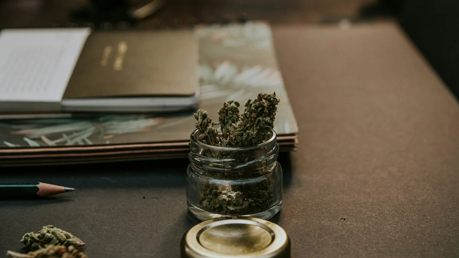 A glass jar of cannabis flower open on a dark desk beside notebooks and a pencil