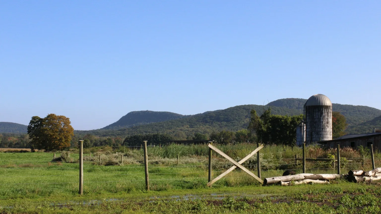 A Western Massachusetts farm with a silo and fenced pasture below a wooded ridgeline on a clear day
