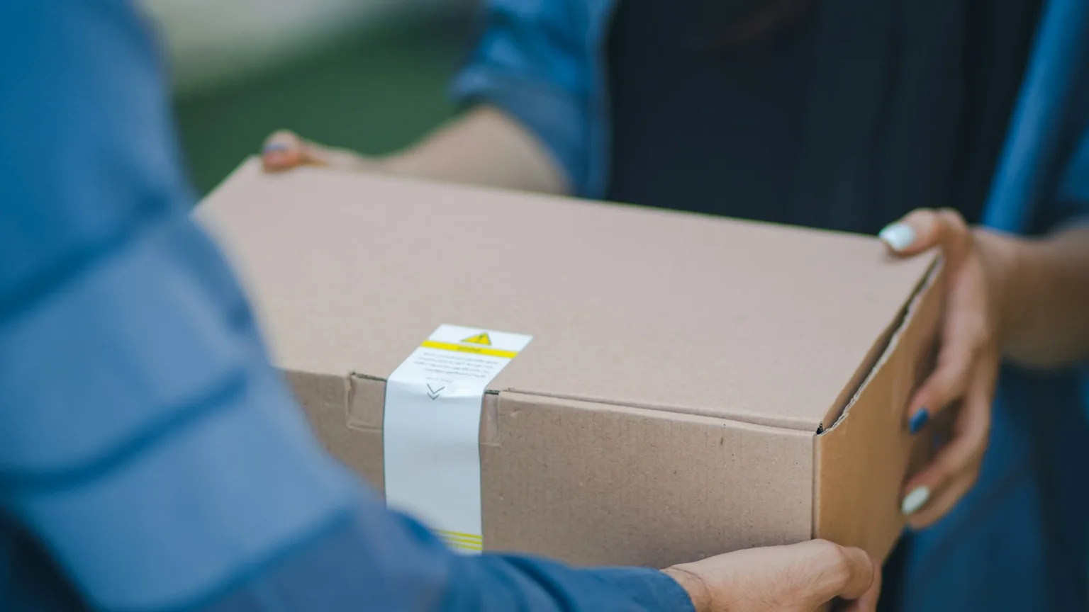 A sealed, unbranded cardboard package with a tamper-evident seal being handed from a delivery agent to a customer