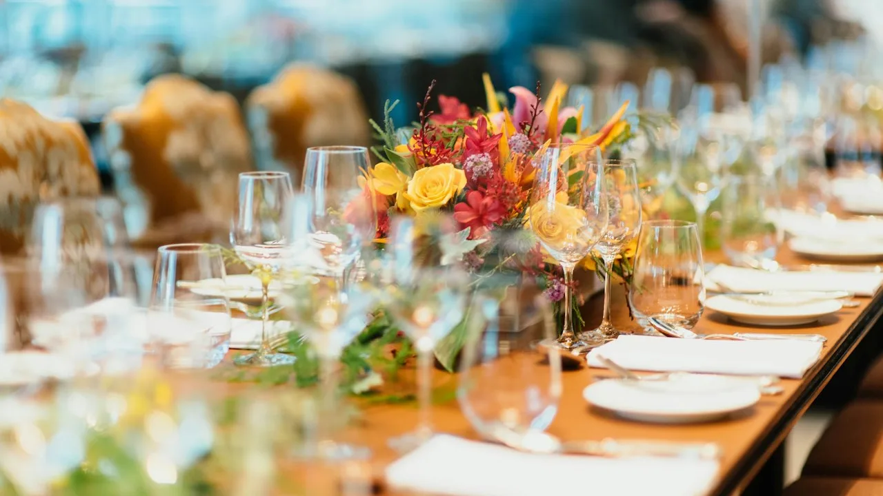 A long dinner table set with wine glasses, napkins, and a bright flower centerpiece